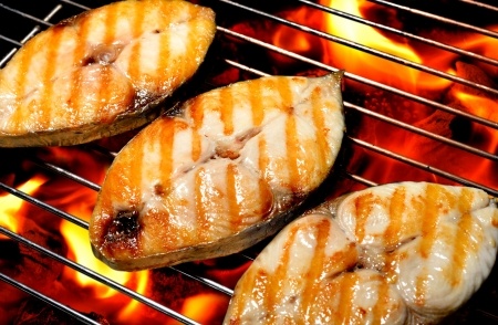 Three pieces of fish steak are being grilled over an open flame, showing visible grill marks on the surface.