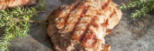 Grilled pieces of meat with grill marks on a stone surface, garnished with sprigs of fresh green herbs.