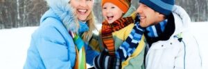 Three people in winter clothing smile outdoors in a snowy landscape with bare trees in the background.