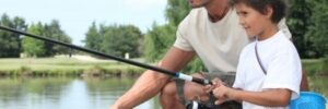 An adult and a child are fishing together by a lakeside, both holding fishing rods and looking at the water.
