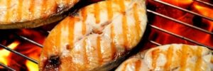 Three pieces of fish with grill marks cooking over an open flame on a metal grill.