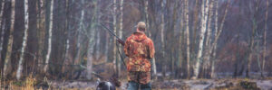 Person in camouflage clothing walking with a dog and holding a rifle in a wet, wooded area during autumn or early spring.