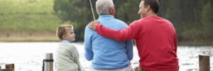 Three people sit on a wooden dock by a lake, with one holding a fishing rod and another with an arm around the third.