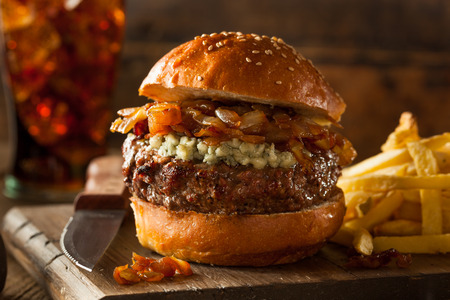 A cheeseburger with caramelized onions and blue cheese on a bun, served with French fries on a wooden board.