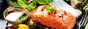 Raw salmon fillet with herbs, sea salt, pepper, tomatoes, olive oil, and vegetables on a dark surface.