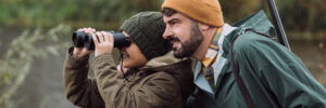 An adult and child in outdoor clothing observe nature by a river; the child uses binoculars, and the adult holds a shotgun.