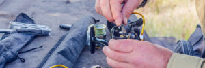 Person assembling a fishing reel at an outdoor table with fishing gear and tools nearby.