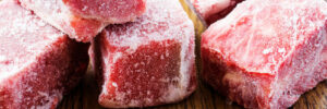 Several frozen raw beef cubes on a wooden surface with scattered salt and peppercorns nearby.