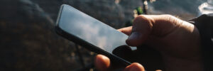 A hand holding a smartphone outdoors near water, with the screen reflecting the sky.