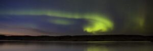 Green aurora borealis lights streak across the night sky, reflected in a calm body of water with distant hills.