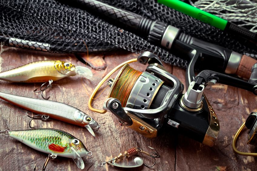 Fishing reel, rod, lures, and hooks on a wooden surface with a black fishing net in the background.
